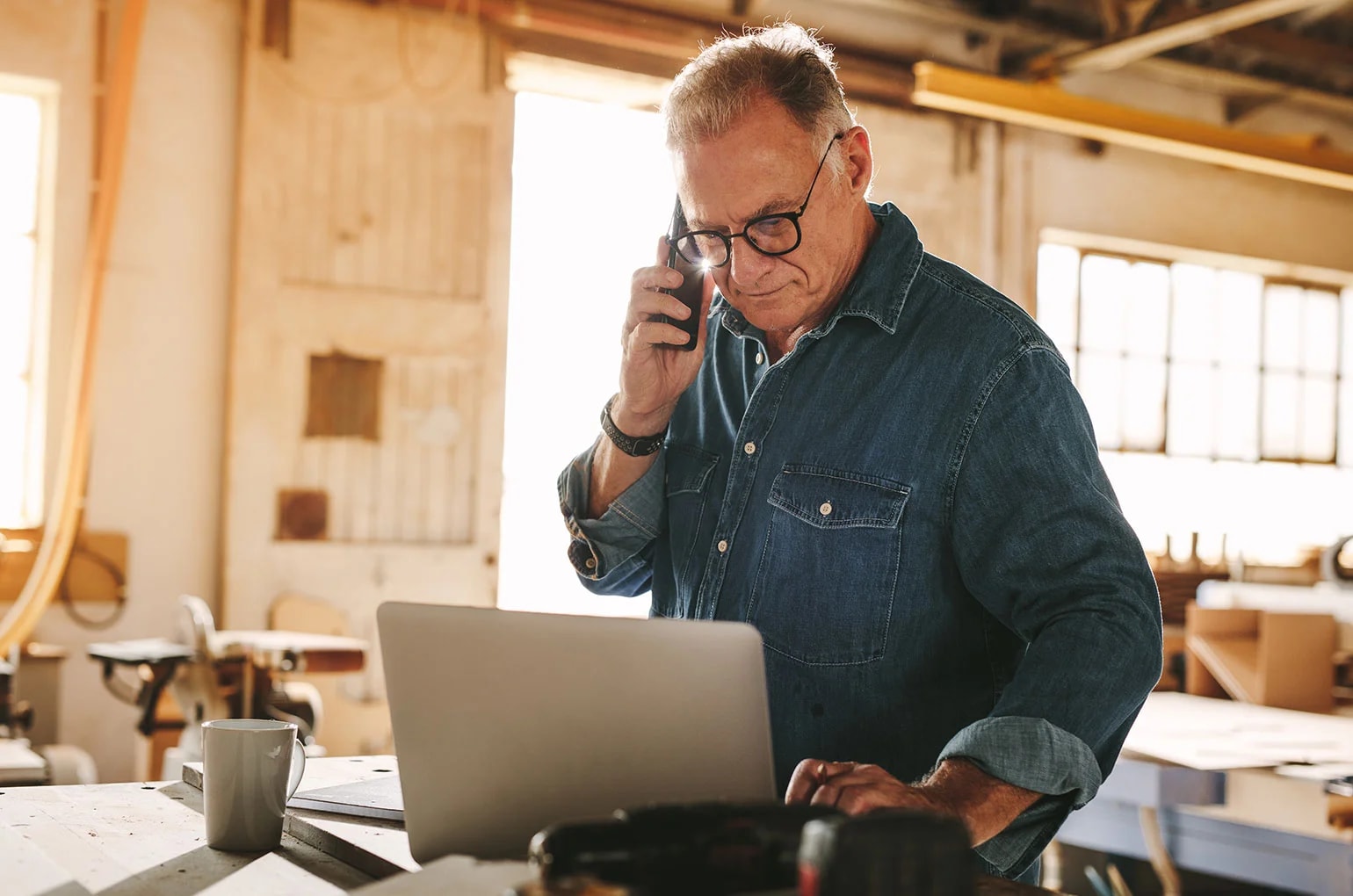 Person arbeitet in einer Werkstatt mit Laptop und Smartphone zwischen Maschinen und Werkzeugen – Symbolbild für Digitalisierung im Mittelstand und den Einsatz digitaler Technologien im Arbeitsalltag.