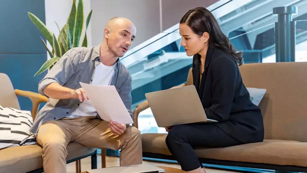 Zwei Personen sitzen in einem modernen Bürobereich und besprechen Inhalte anhand von Dokumenten und einem Laptop.