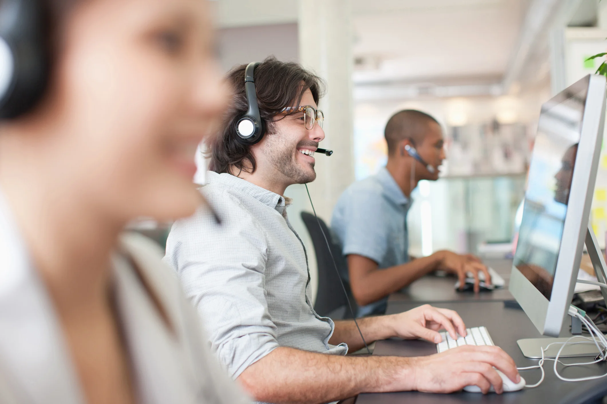 Mehrere Personen sitzen nebeneinander an Computern und tragen Headsets; Fokus auf eine Person im Vordergrund beim Tippen, modernes Büro im Hintergrund