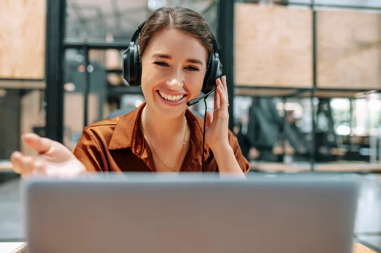 Person mit Headset arbeitet am Laptop in einem modernen Büro mit Glaswänden; eine Hand am Headset, die andere nach vorne gestreckt.