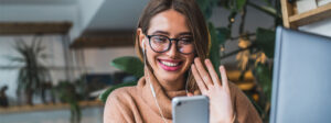 Das Bild zeigt eine Freelancerin in einem Café, die gerade winkend in ihr Smartphone schaut.