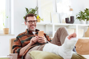 Mann mit Gips am Bein liegt auf der Couch und lächelt in sein Handy