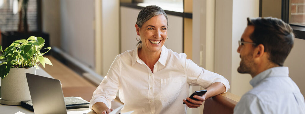 Frau spricht lächelnd mit Mann an einem Schreibtisch vor einem Notizbuch