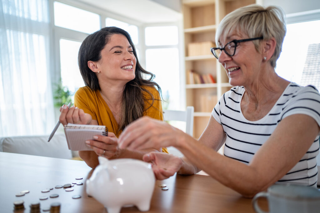 Eine jüngere und eine ältere Frau sitzen an einem Tisch, vor ihnen steht ein Sparschwein. Sie unterhalten sich über finanzielle Planung und Altersvorsorge für Selbständige.